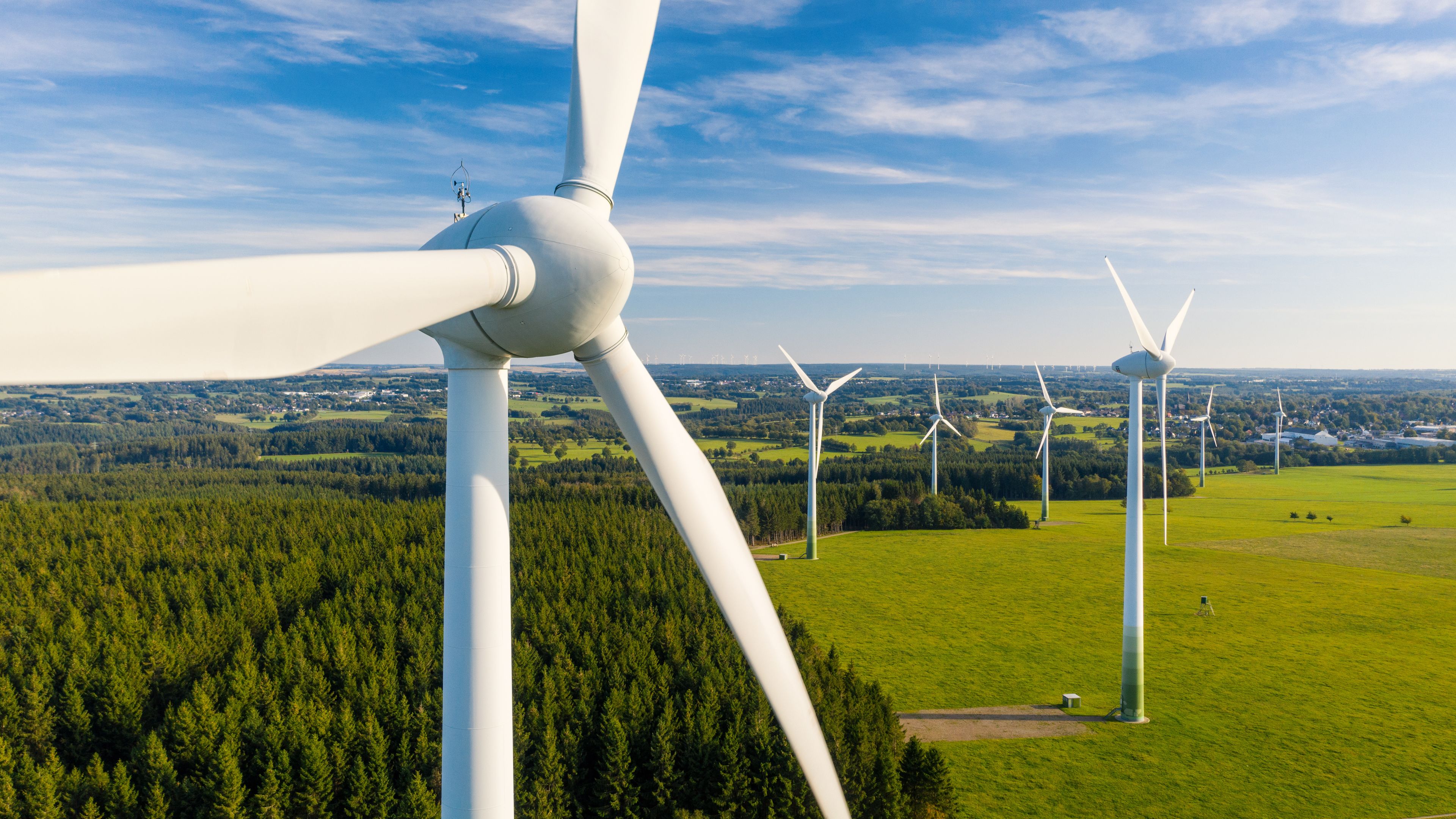 A scenic view of a wind farm with multiple white wind turbines standing tall in a grassy field surrounded by dense forests. The sky is clear and bright blue with scattered clouds. The landscape stretches into the distance, showcasing green fields and distant trees.