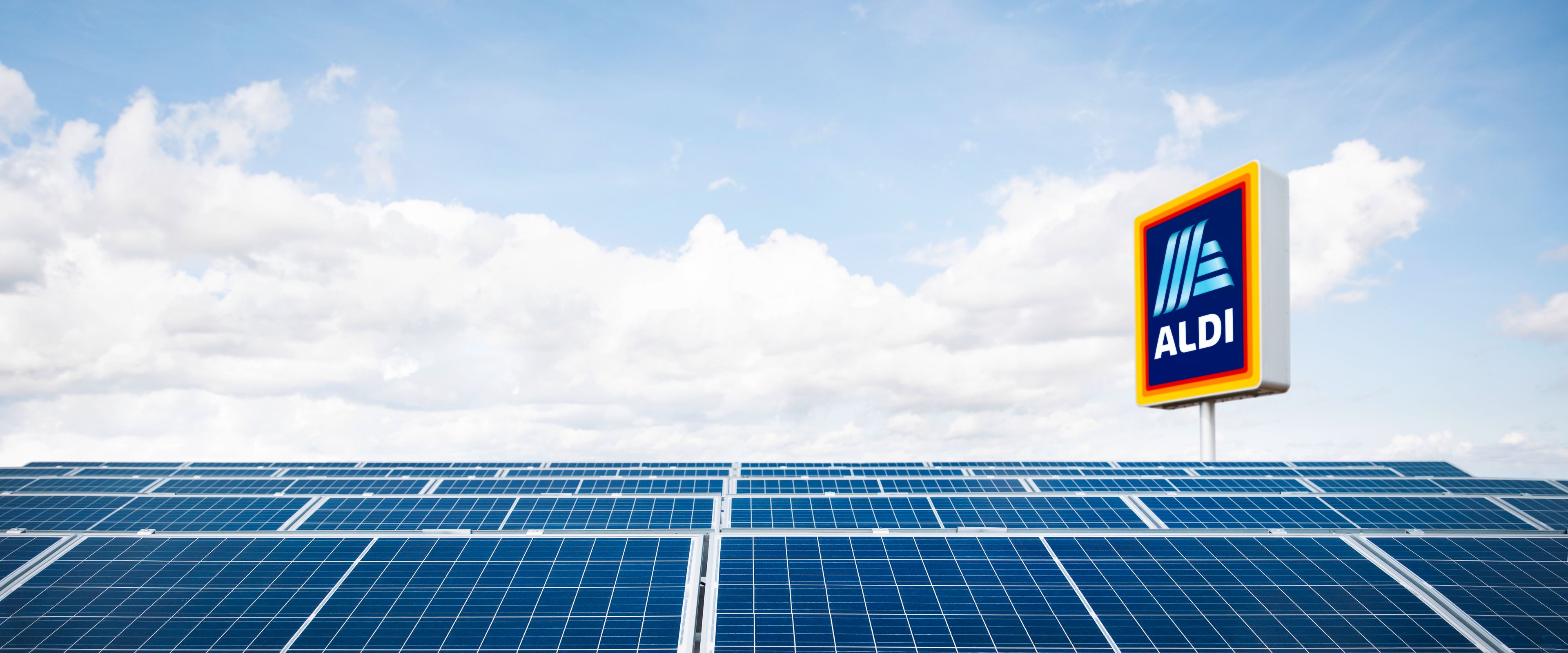 A solar panel array under a bright blue sky with scattered clouds. In the background, a sign with the Aldi logo is visible, indicating the location of the panels near an Aldi store.